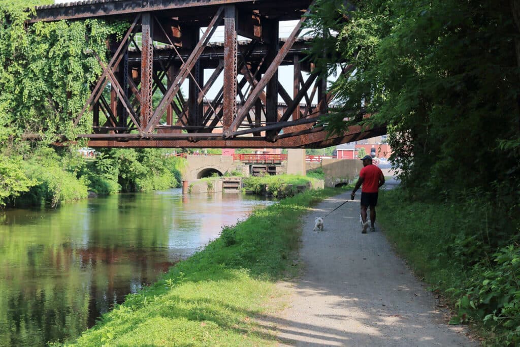 Person walking a small dog on the gravel Delaware Canal Towpath beneath an old steel railroad bridge