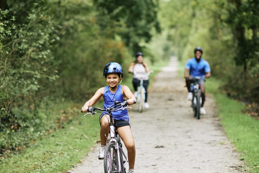 Girl in a bike helmet riding toward the camera on the crushed-stone NCR Trail, with two riders behind her