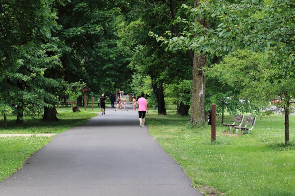 People walking a paved park path lined with benches and mature shade trees at Neshaminy State Park