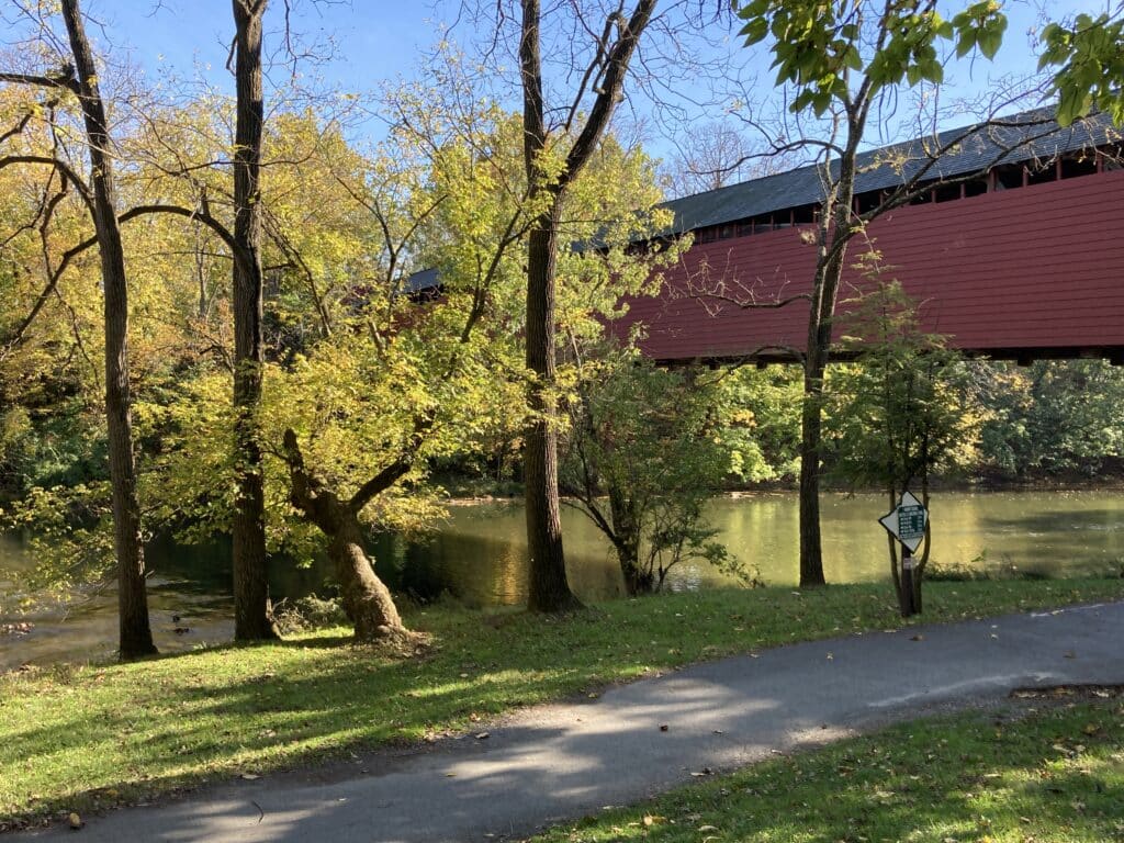 Paved trail beside a creek in early fall, with a red covered bridge just off the path